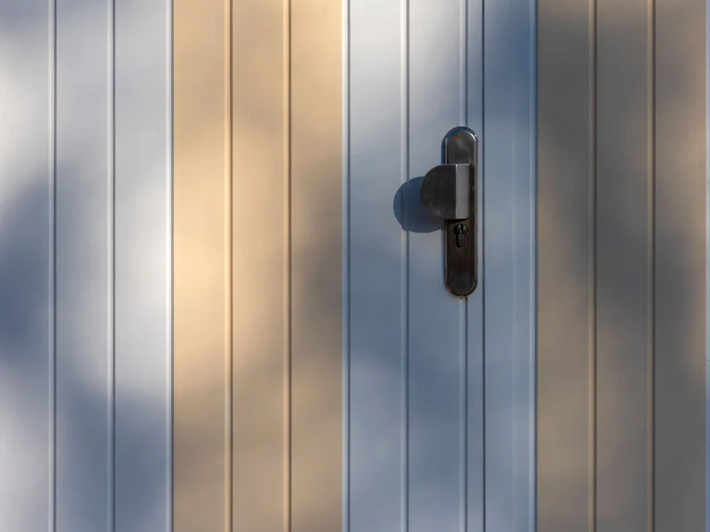 Vertical aluminium panel door with a lever handle, low sunlight raking across the surface